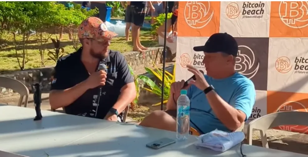 Pedro Vital conversando en un evento con backdrop de Bitcoin Beach, El Salvador