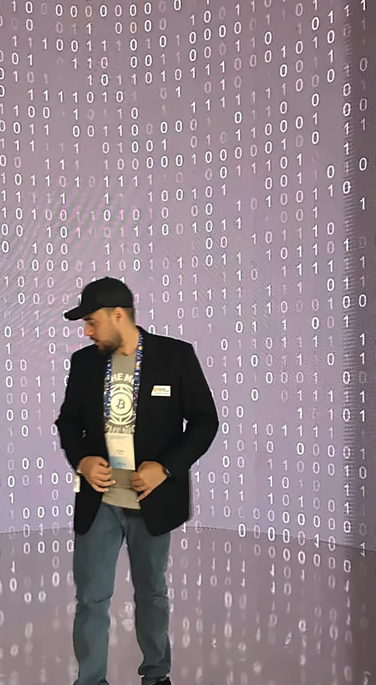 Pedro Vital de pie frente a una pared cubierta de código binario, con la mirada baja en actitud reflexiva.
